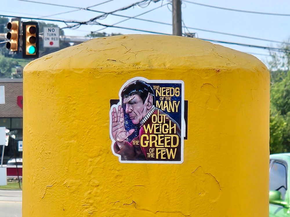 A cement bollard painted yellow. There's a sticker on it with a drawing of Spock giving a Vulcan salute, and it says "the needs of the many outweigh the greed of the few."