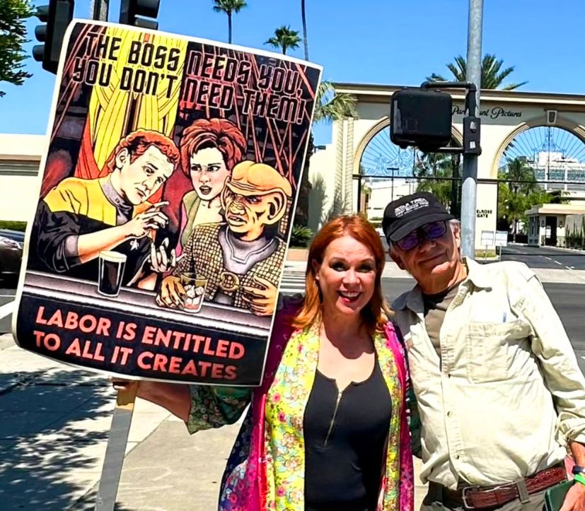 Chase Masterson, who played Leeta, carrying a poster-sized print of the same image attached to a picket during the WGA SAG-AFTRA strike in front of Paramount Studios. She is posing for a picture with a man I don't recognize.