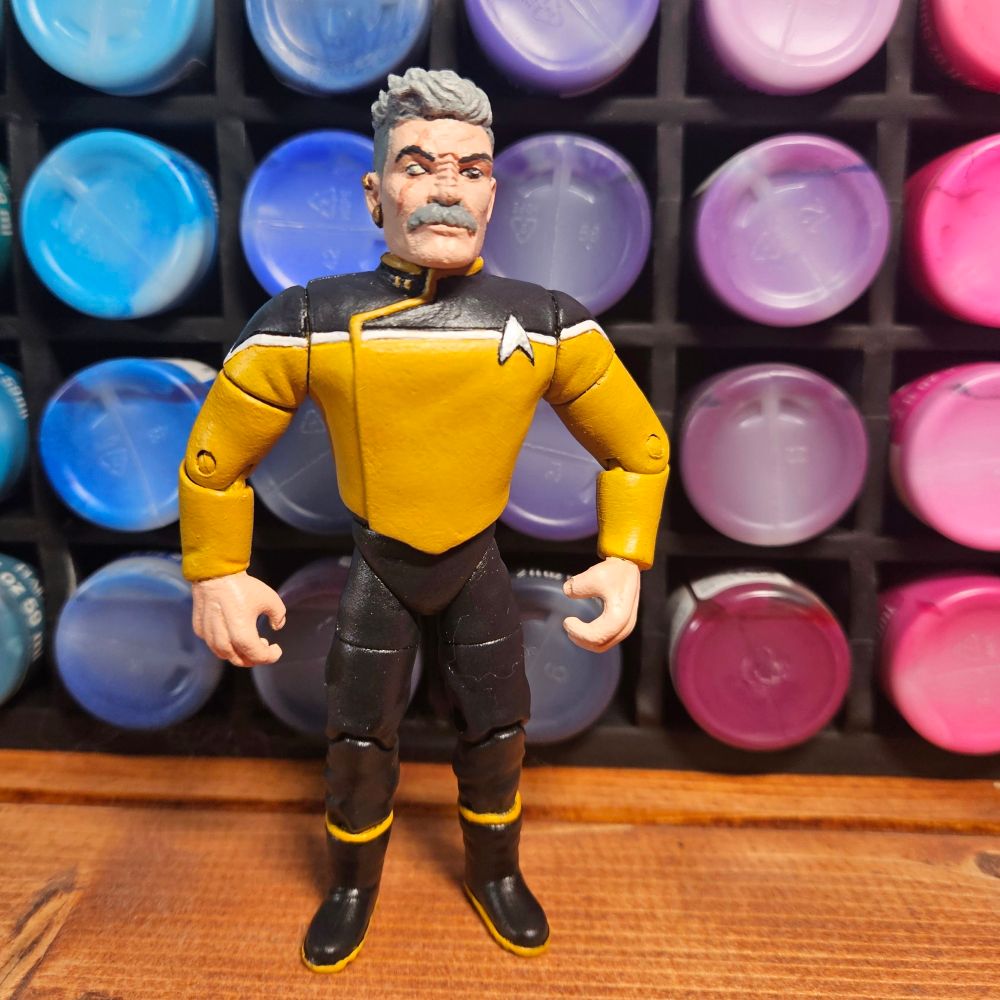 A custom action figure of Lieutenant Shaxs from Lower Decks in front of a rack of acrylic paints.