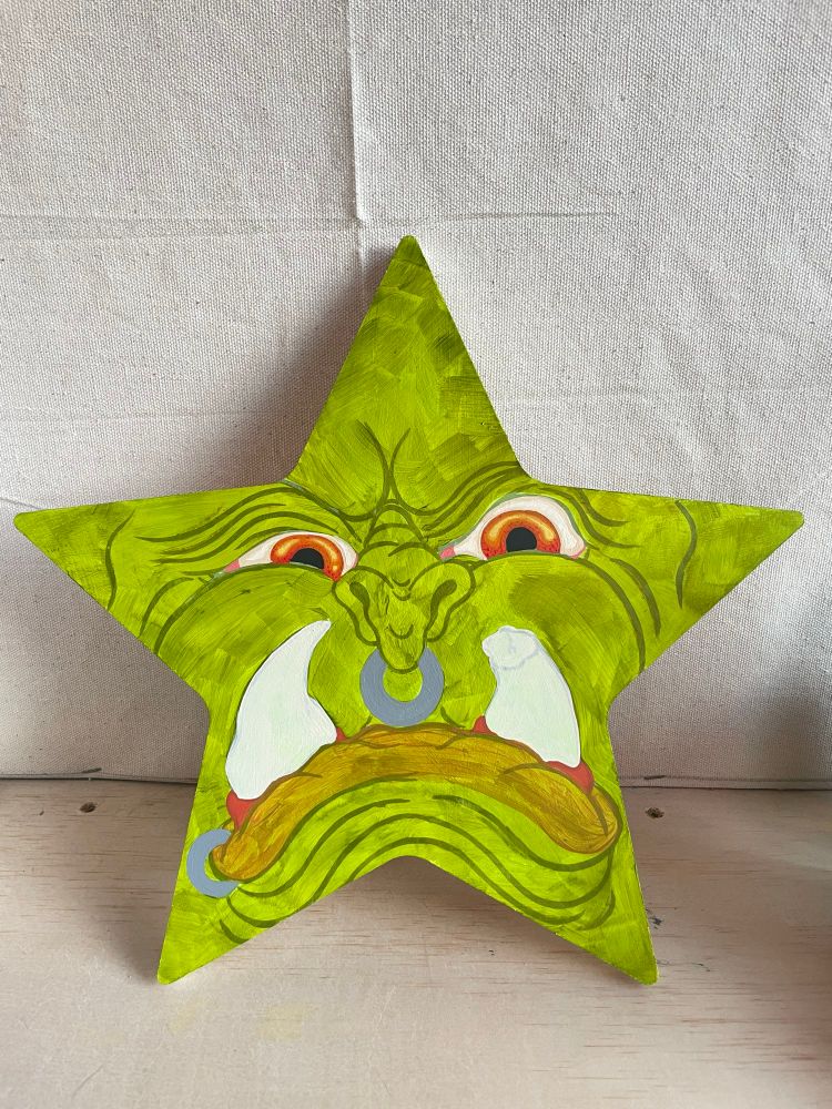 Photo of a star shaped canvas with a green orc roughly painted in. He has two huge tusk like teeth, a large septum piercing and lip piercing. His eyes are painted with more detail than the rest of him. The eyes are orange, yellow, and a bit of red 
#art #fantasyart #acrylicpainting #monsterart #monster #orc 