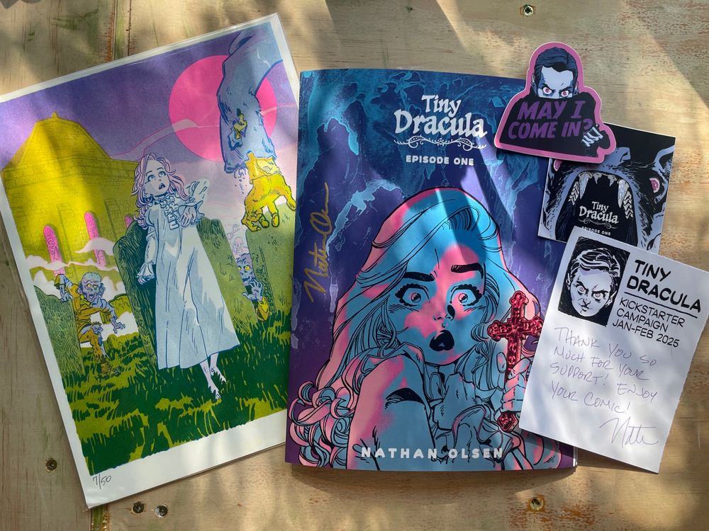 Photo of a spread of art including a riso print with young woman Lucy walking through a zombie infested grave yard, a comic book with the same woman on the cover, a sticker of Dracula saying “May I come in?”, and a thank you note from a kickstarter campaign 