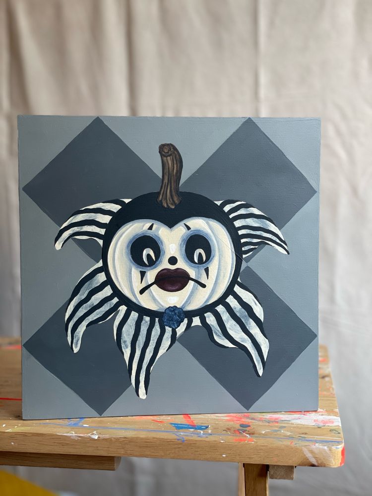 Close up picture of the painting of a sad clown pumpkin. Shes a white pumpkin, with dark circles around her cut out eyes, and a dirty white and black striped collar around her pumpkin head 