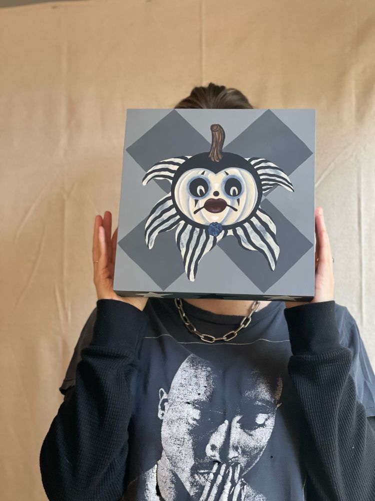 Human holding a square painting up of a sad clown with a black hood and dark red lips 
