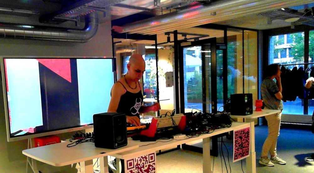 CyberGwen in concert, standing behind a standing desk full of synths and other gear, with a visualizer on a big screen behind her.