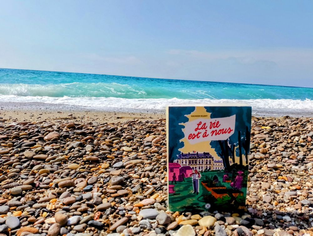 Livre "La vie est à nous" posé sur les galets devant la Méditerranée sous le ciel bleu 