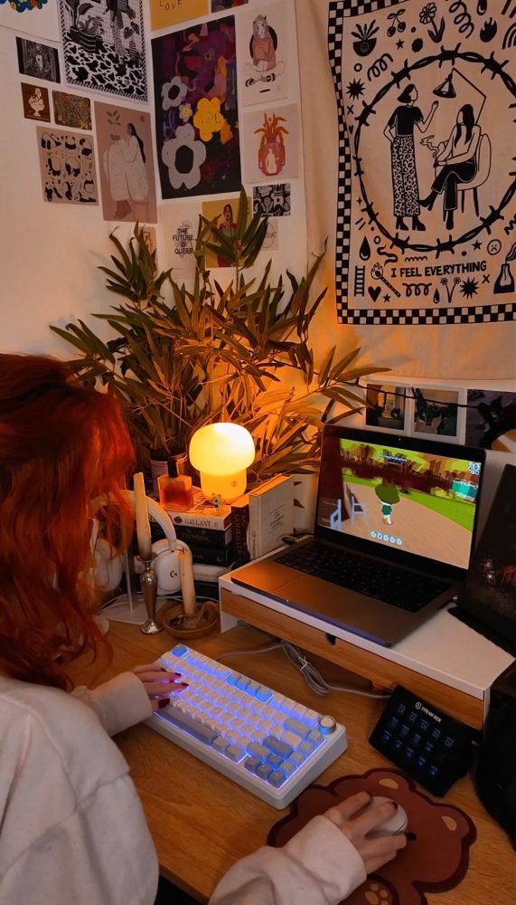 A picture of honey's desk setup. Honey is looking away from the camera at their laptop on the desk, their keyboard is white and blue and Webfishing is being played on the laptop. There is a cozy lamp in the corner of the desk shining very orange sitting on top of a book stack. There is a large fake plant in the corner of the desk. There is a tapestry and posters on the wall of art