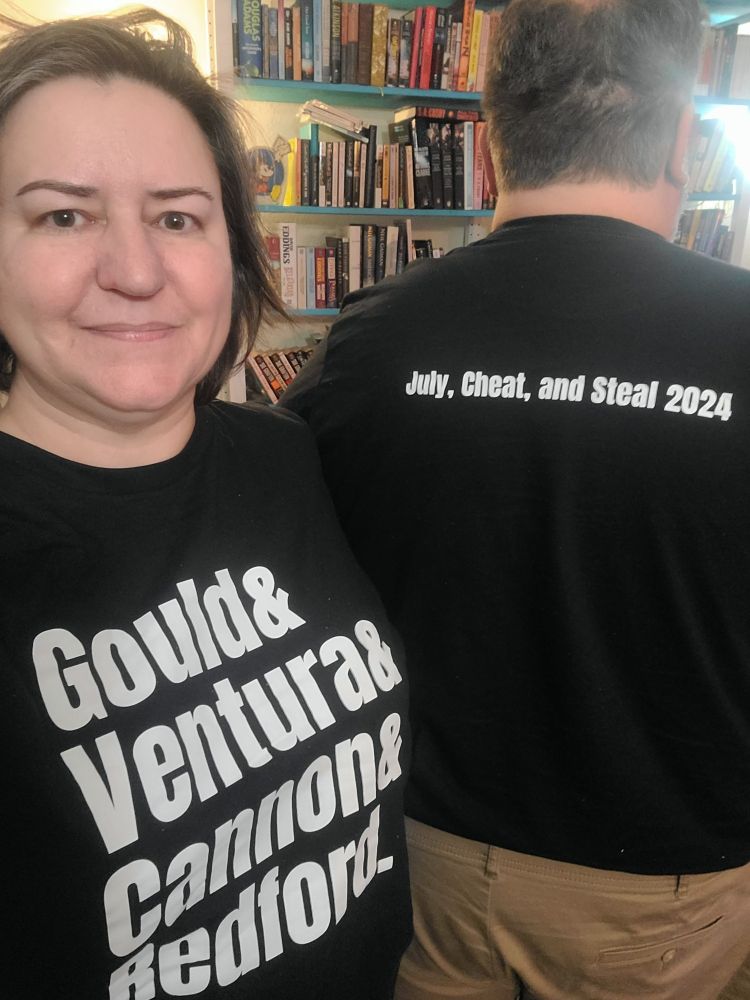 Me on the left, facing the camera. I'm wearing a black shirt with white lettering that reads 
Gould&
Ventura&
Cannon&
Redford.
My husband is to the right of frame, facing away from the camera, the back of his black shirt reads in white lettering "July, Cheat, and Steal 2024"