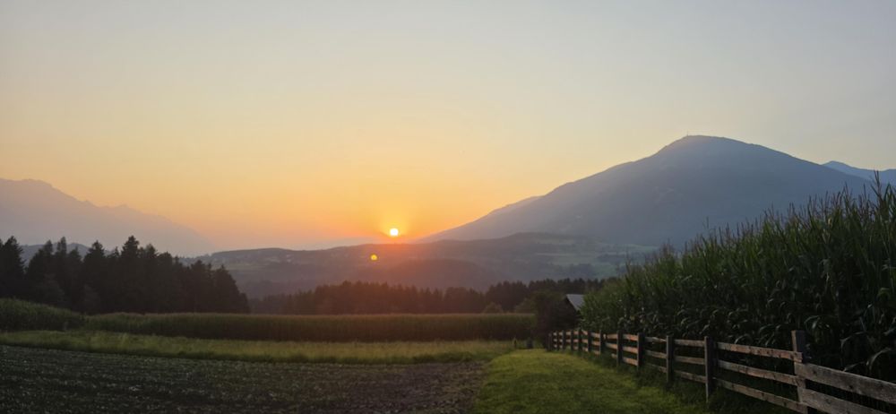 Sonnenaufgang am Horziont mit orang/rot leuchtendee Sonne.
Berge & Felder