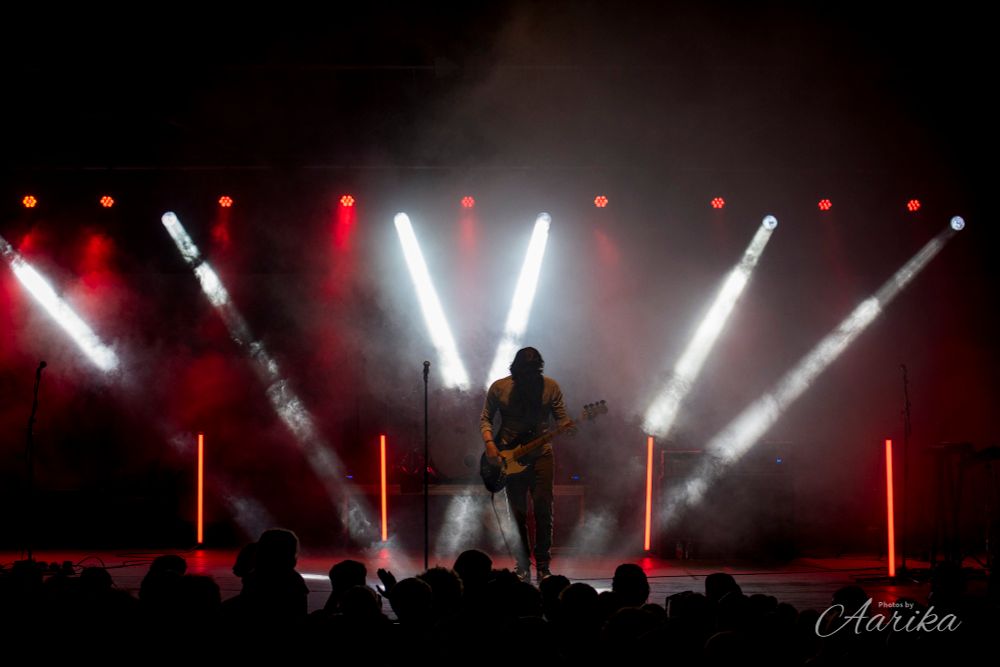 Bassist Ben Spivak of MAGIC! stands playing a solo in a partial silhouette surrounded by red and white lights. 