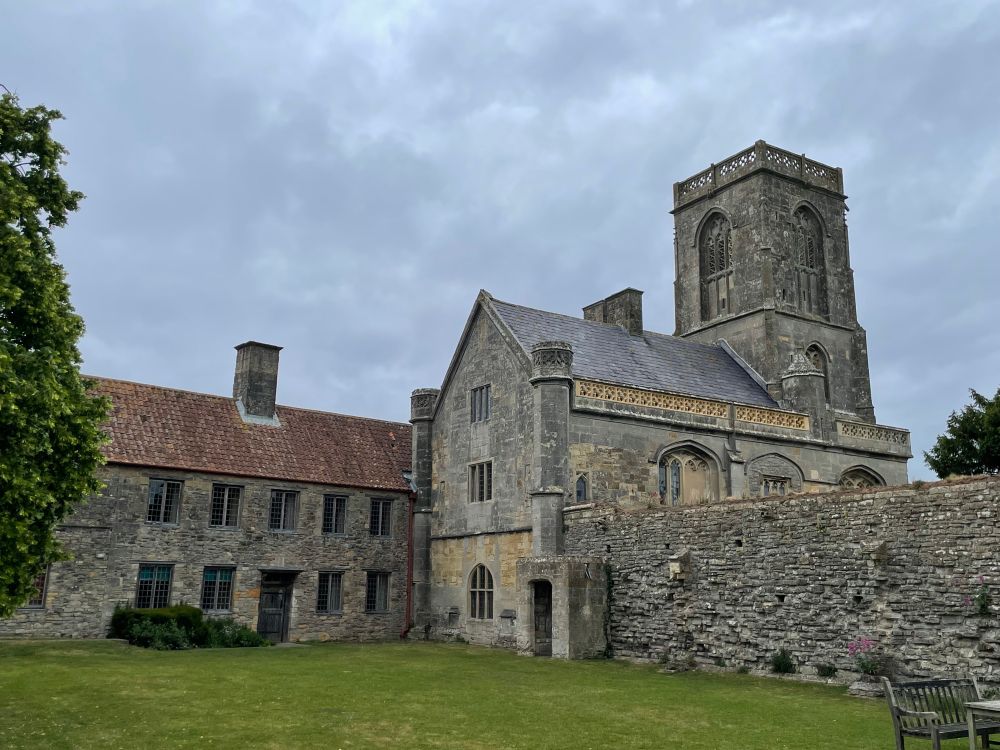 View of the Priory, with the nave, crossing tower, and remains of the cloister wall on right, and later, lower range (former farmhouse) attached to NW corner of nave. Greyish sky, green lawn, tree to left edge of view.