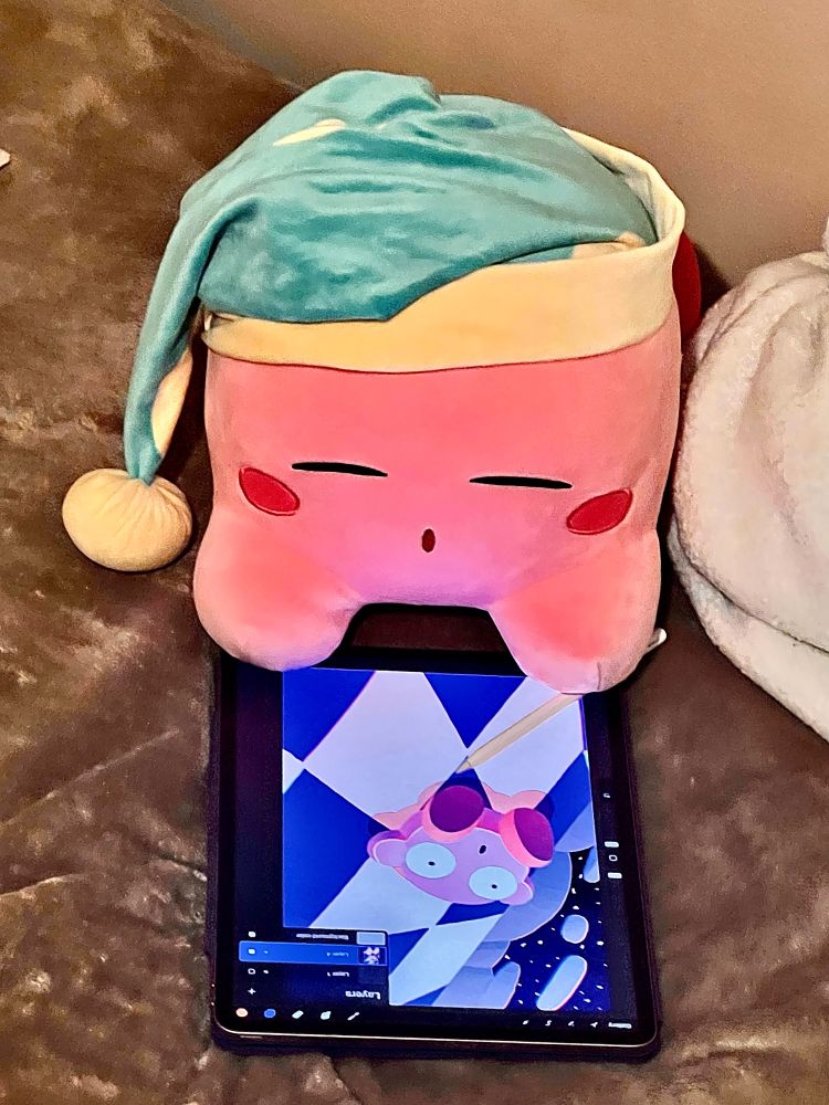 Kirby sleepy plush set up on a drawing tablet to make it look like it’s drawing on it! The tablet shows a work-in-progress drawing of Kirby sliding down a slope. 
