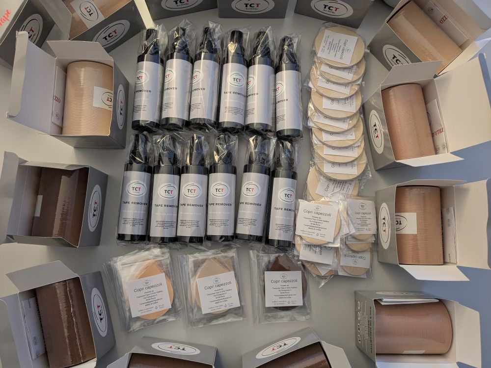 A pile of more than a dozen trans tape in three different skin colored hues, nipple stickers in the same hues and 12 bottles with oil based trans tape remover