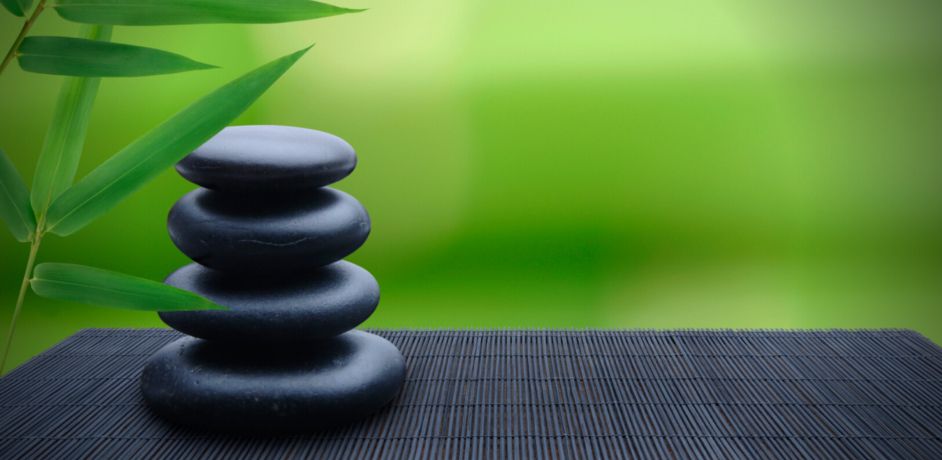 Zu sehen sind hier 4 schwarze runde Steine zu einem Turm zusammengesetzt auf einer schwarzen Matte. Im Hintergrund ist ein grüner Zweig und verschwommen grüne Landschaft.