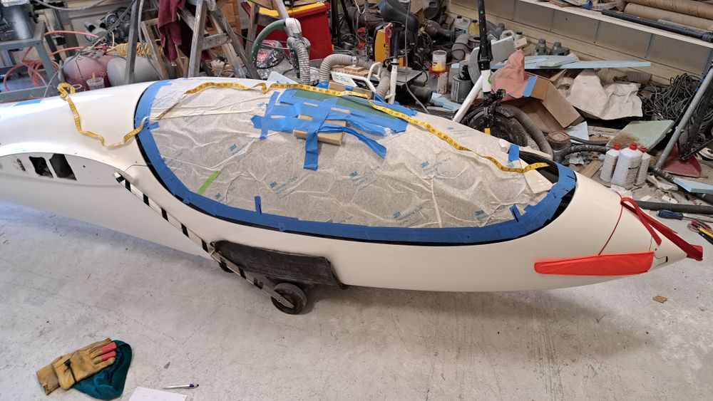 A yellow tape measure lies along the top of the glider's canopy. The canopy is covered with protective paper and edged with blue tape. The fuselage nose is at the right edge of the frame, which has two folding props covered in bright orange fabric.

The garage floor is gray and behind the fuselage is the clutter of various equipment and sundry items.