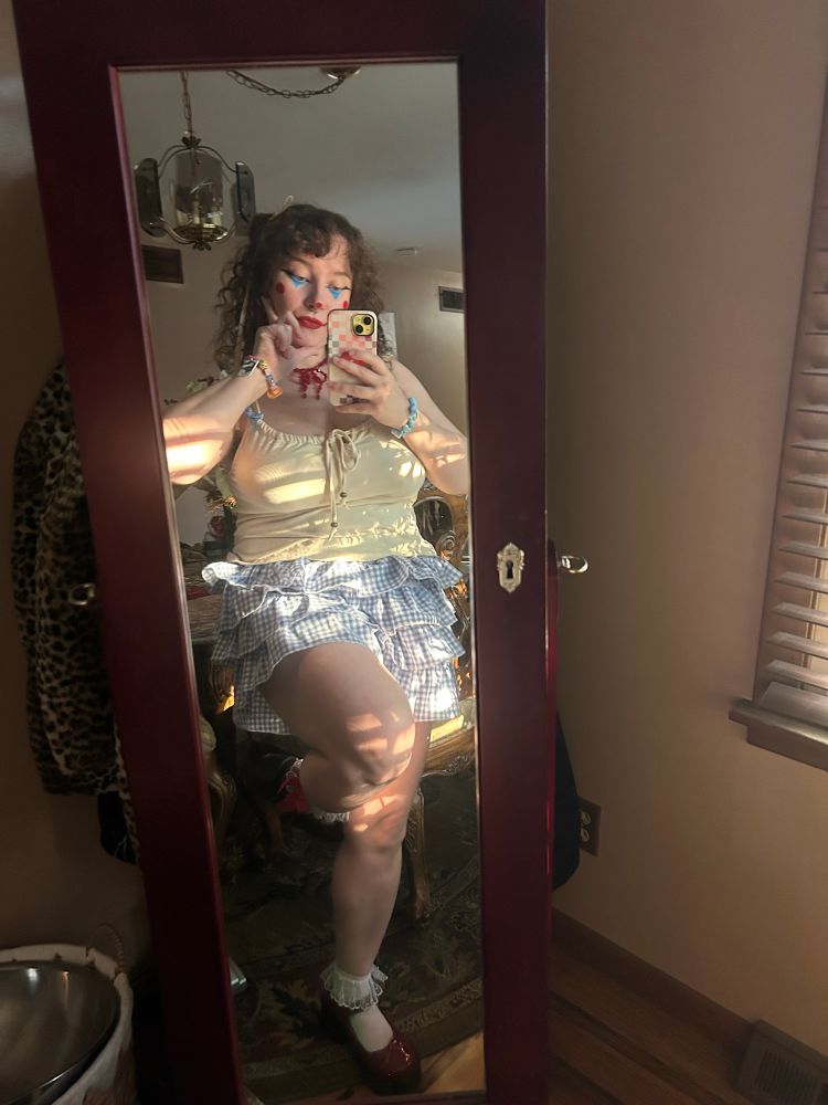 pic of me with clown makeup, leg up, smiling, red bow necklace, gingham baby blue and white bloomers, frilly white socks, red mary janes, light yellow tank top  