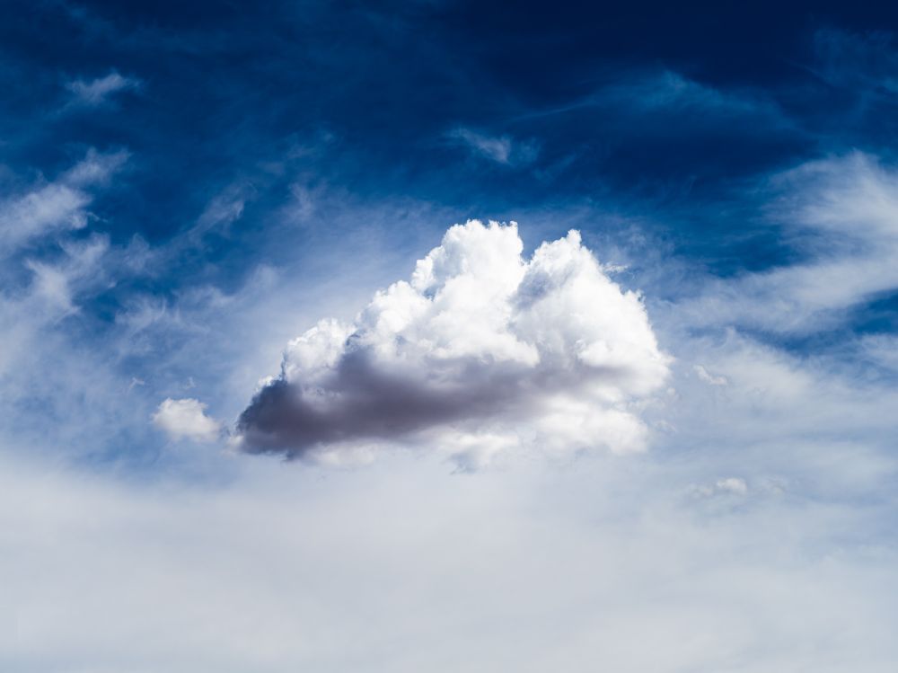 a single fluffy cloud with a purple-grey underbelly floats alone in the sky. the top half of the sky is deep blue with wispy cirrus clouds. the bottom half is almost solid white with cirrostratus.