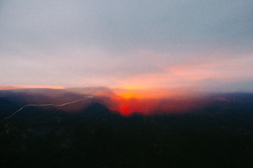 a photograph that looks like an abstract watercolor of a landscape at sunset, velvety blacks of the ground turning into overlapping purple waves of mountains with a lava-red and orange and yellow wash in the center leading to soft pink and blue sky. a single hard white line zig zags from the left of the frame towards the sunset
