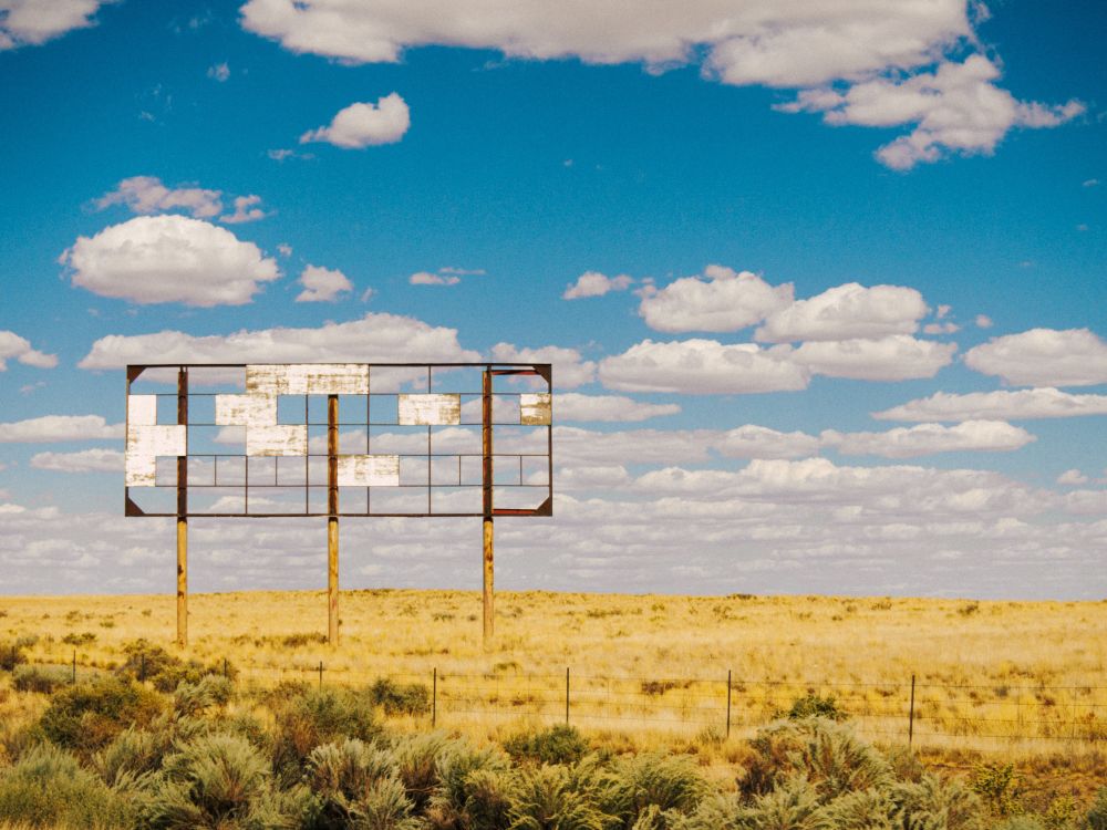 a billboard stands in a yellow desert field under a perfect blue sky. but the billboard is very old, and it used to have a plywood sign on it, but the sign is almost entirely gone, with just random squares of weathered white wood remaining 