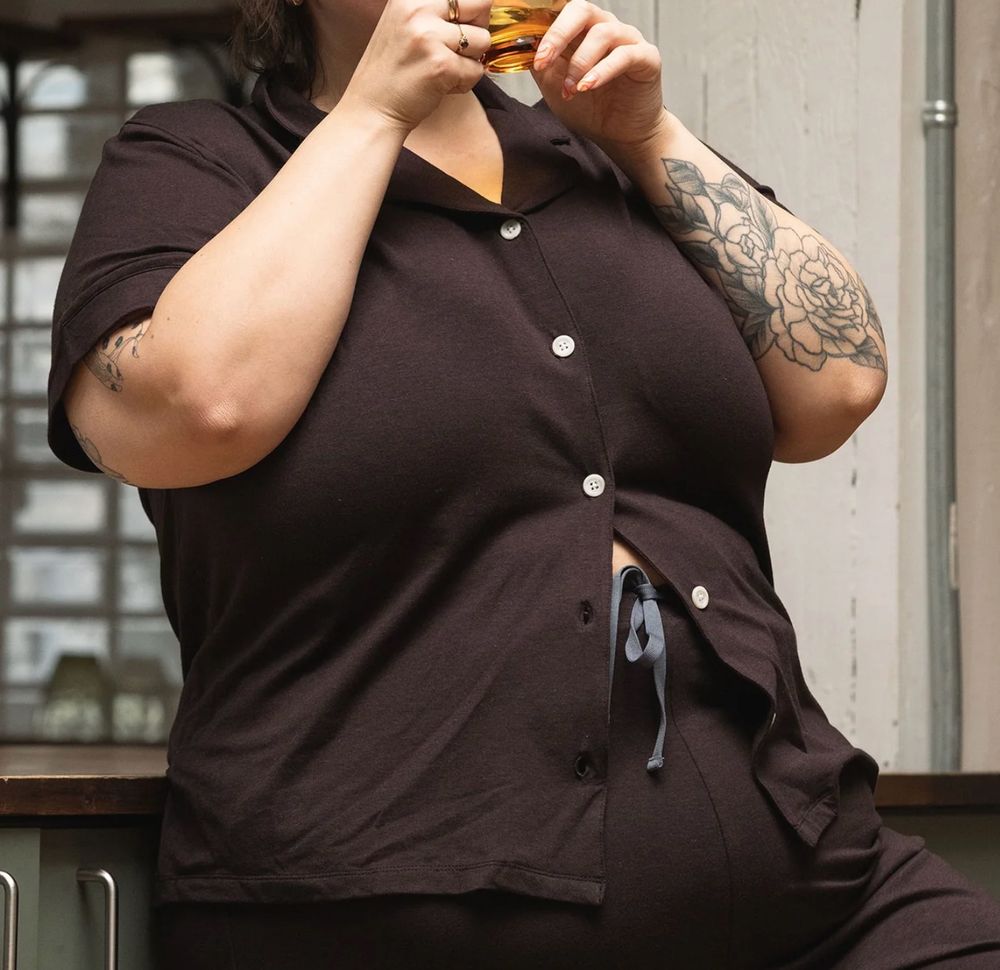 crop of plus-size wearing a shit brown pajama (?) top that has large, wide spaced white buttons and a childish flat collar — granny wear strikes again — and the bottom 2 buttons under her breast are unbuttoned, because THE SHIRT DOESN'T FIT HER STOMACH

also it's wrinkly! they didn't iron it!