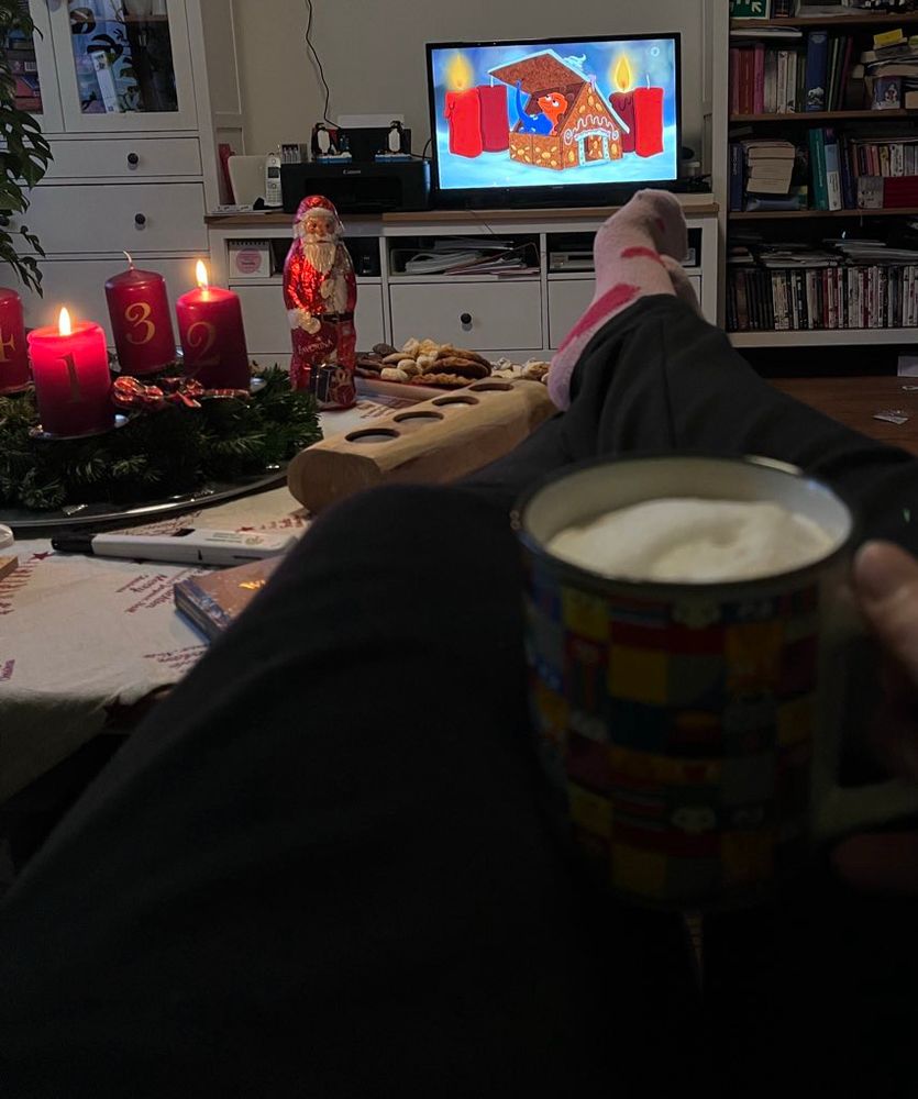 Kaffee, Beine auf Couchtisch, Adventskranz mit 2 brennenden Kerzen, im Hintergrund der Fernseher mit dem Intro der Sendung mit der Maus.