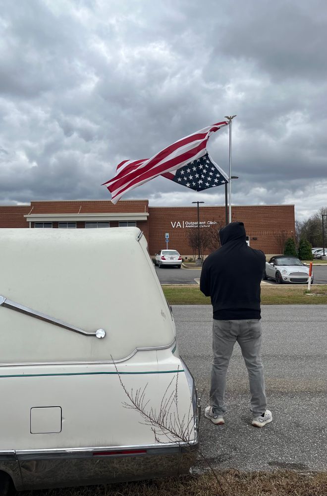 Veteran Army Sgt. Leland Fullbright stand holding an upside down American flag next to his white Cadillac hearse. His back is to the photographer. The Anniston-Oxford VA is in the background. 
