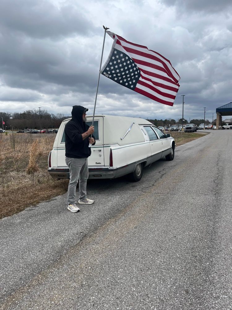 Veteran Army Sgt. Leland Fullbright stand holding an upside down flag next to his white Cadillac hearse. 
