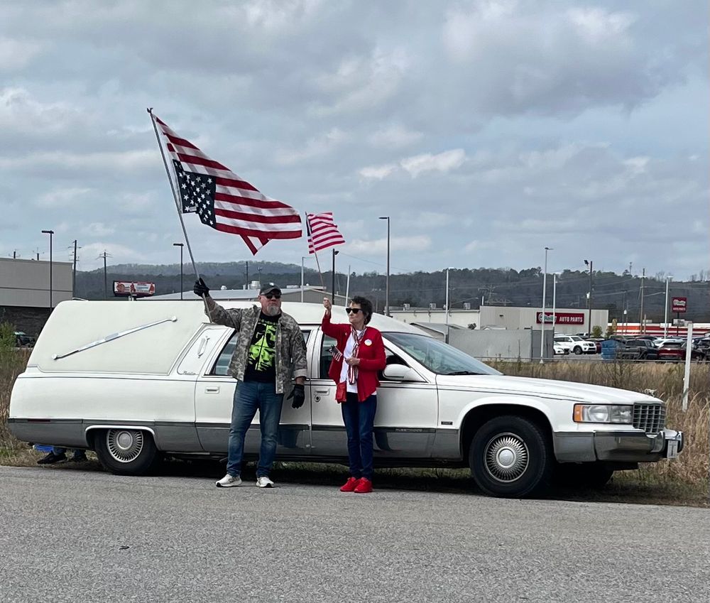Army Veteran Sgt Leland Fullbright and another hold American flags in front of his white hearse. 
