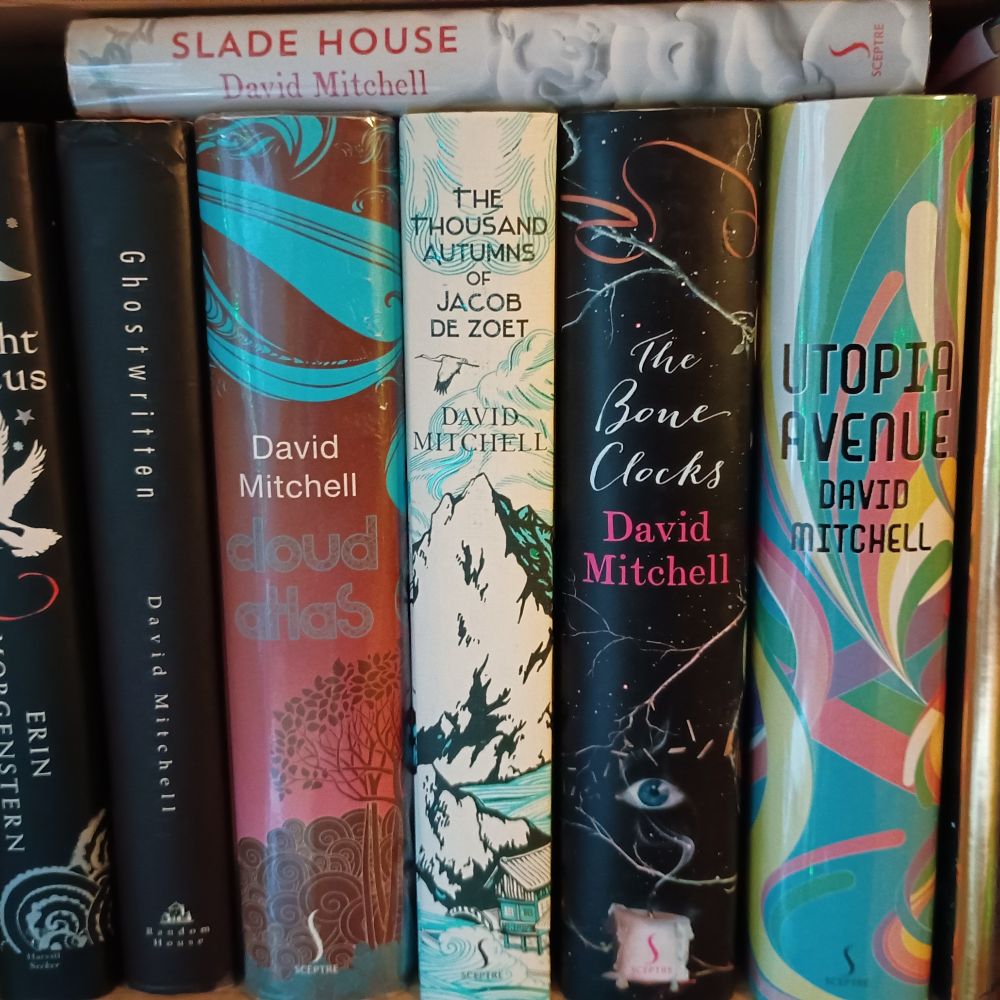 Selection of David Mitchell books, spines out, on a bookshelf. Left to right: Ghostwritten, Cloud Atlas, The Thousand Autumns of Jacob De Zoet, The Bone Clocks, Utopia Avenue, with Slade House sitting flat atop these.
