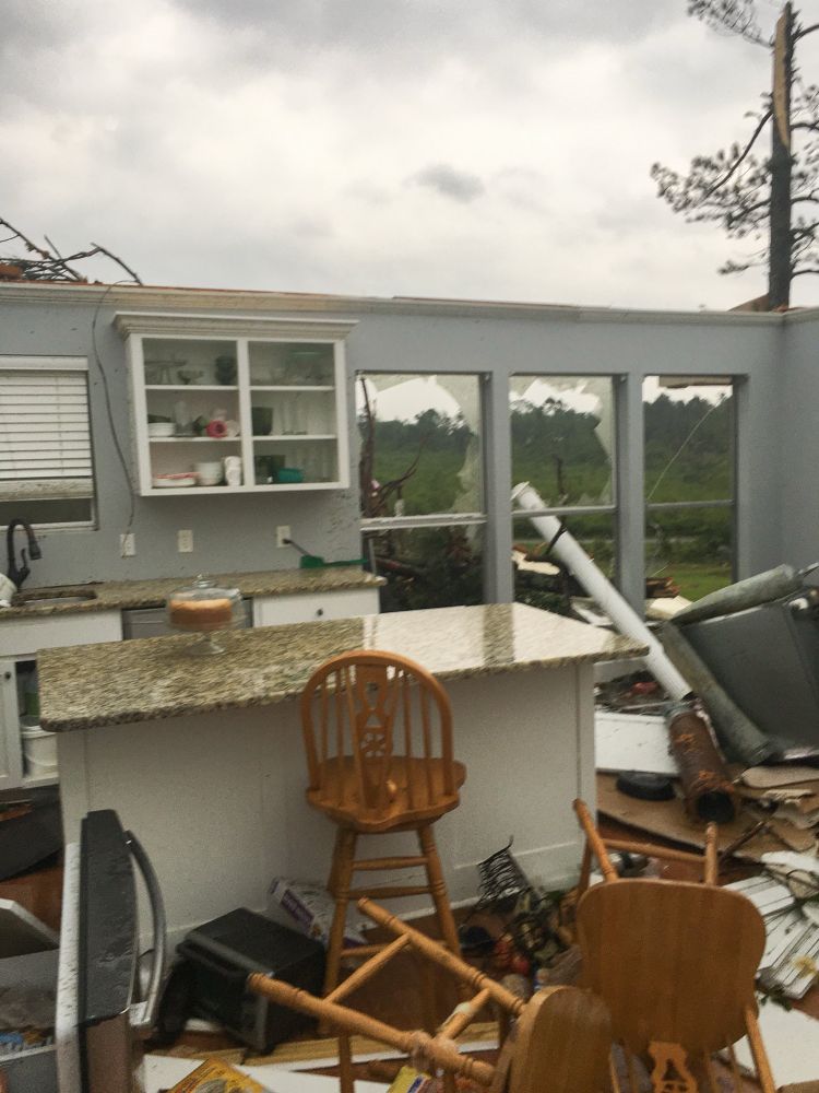 A disheveled and mostly destroyed suburban home sits without a roof or windows in the aftermath of a violent tornado, but on the kitchen island sits a cake in a glass cake dish as if the chaos around it never happened