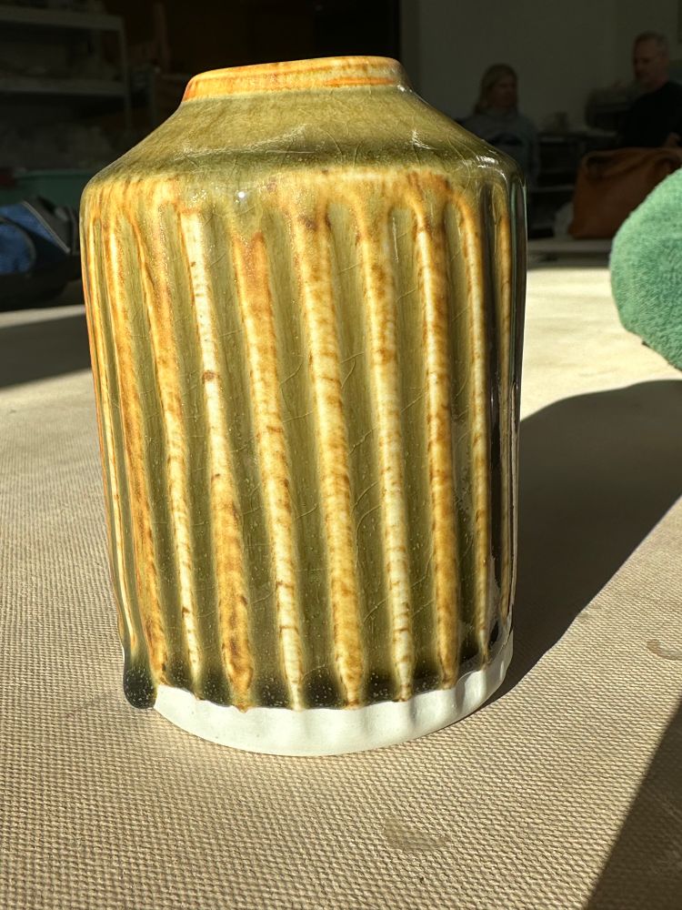 A small porcelain vase with facets carved, with the same glaze as the cup. The glaze is a transparent dark green where it pools in the facets, and breaks amber with brown spots over the facets. The glaze is called frog belly.