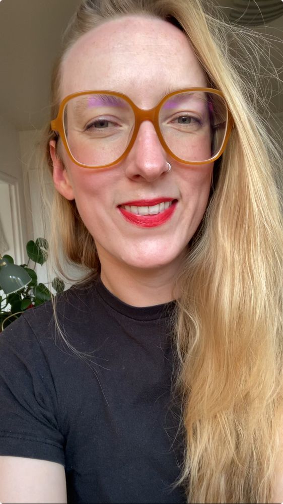 Sage smiles, looking directly at the camera with big glasses and a bright red lip