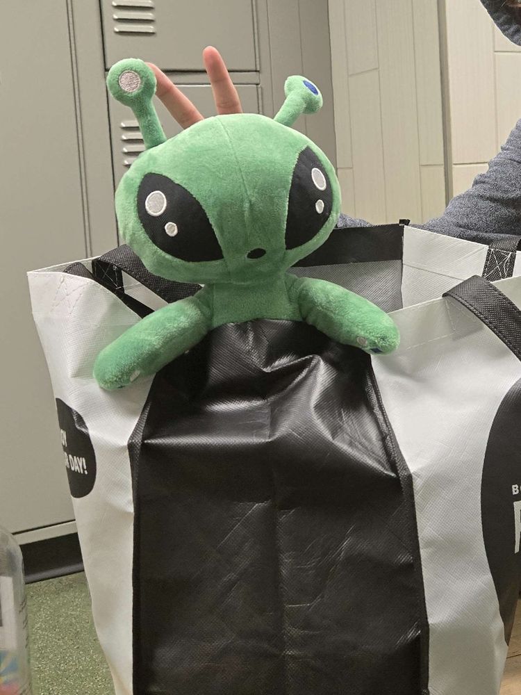 IKEA alien plushie in a reusable bag in a locker room. An arm is reaching behind him giving him bunny ears.