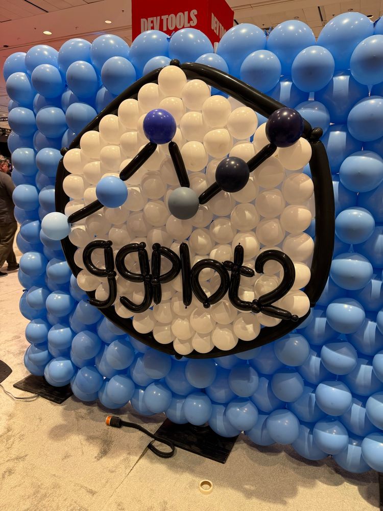 Balloon wall art inspired by the ggplot2 hex sticker. The display features a hexagonal frame made of black balloons, filled with white balloons inside and surrounded by a background of light and dark blue balloons. A line graph with black balloon segments and round coloured nodes runs across the centre, and the word “ggplot2” is spelled out in black balloon letters below the chart.