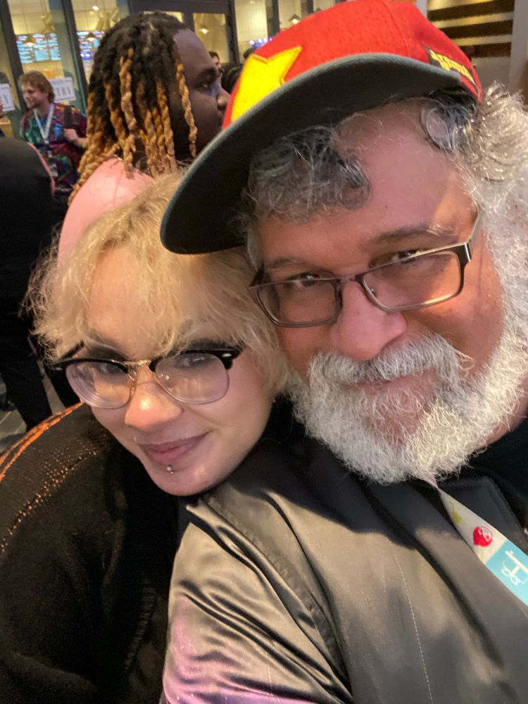 Two beautiful mixed race folks one femme and the other masculine. They are wearing glasses and hugging and smiling at the camera. 