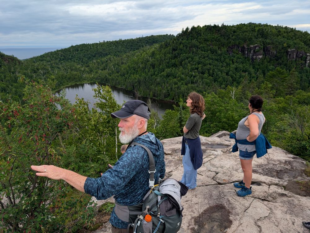 A man in hiking equipment points in the distance, while two educators stand on a rocky overlook beside him. Credit: MNSG.
