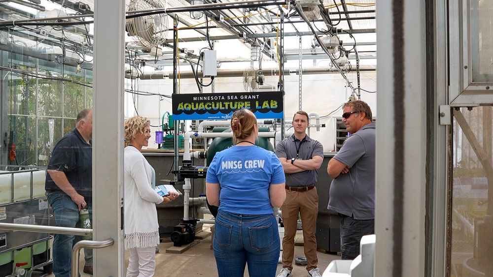 Julia Grenn, MNSG aquaculture extension educator, speaks with a group of legislators and producers in a greenhouse in front of a sign reading "Minnesota Sea Grant Aquaculture Lab." Credit: C. Dettmann/MNSG.