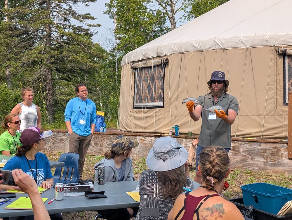 A man in orange gloves holds up two fish in his hands to a crowd of seated educators in front of a yurt. Credit: MNSG.