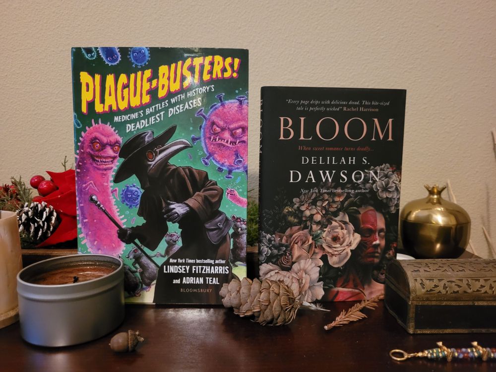 Photograph of 2 books—Plague-Busters by Dr. Lindsey Fitzharris and Bloom by Delilah S. Dawson—on a side table with a candle tin, small box with brass filigree and a brass pomegranate.