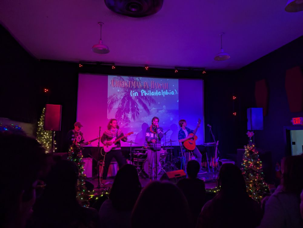 A photo of band, Tucked In, with two guitarists, one bassist, a lead singer, and a drummer performing on a small stage surrounded by holiday decorations and in front of a visual with a palm tree that says “Christmas in Hawaii (in Philadelphia)”