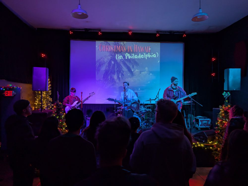 A photo of band, Wallace, with one guitarist, one bassist, a lead singer (also playing guitar), and a drummer performing on a small stage surrounded by holiday decorations and in front of a visual with a palm tree that says “Christmas in Hawaii (in Philadelphia)”