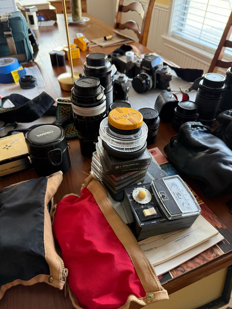 A dining table with stacks of filters and camera lens of all sizes, a vintage light meter and a Nikon F2. 