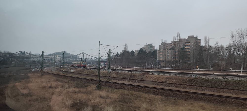 train tracks departing Berlin Gesundbrunnen northbound, with an S-Bahn train in the foreground, some Plattenbau apartments opposite, and bridges and rail infrastructure in the surroundings.
