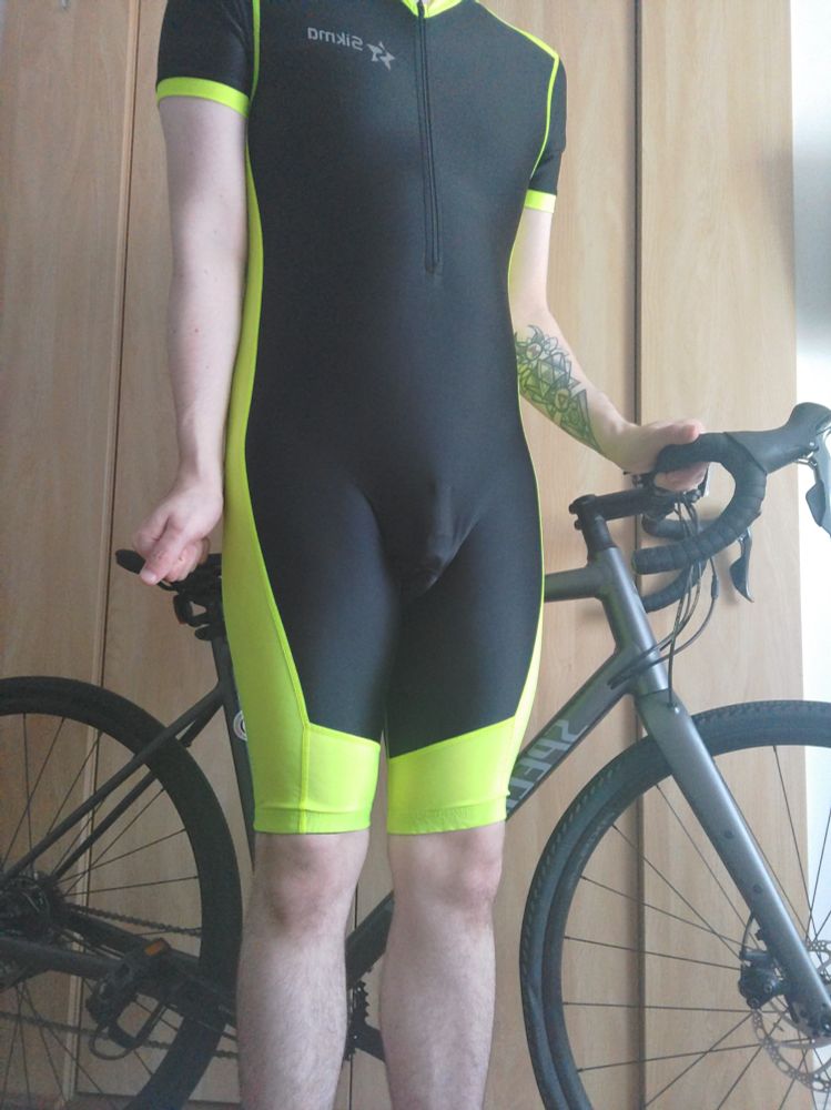 Myself wearing a black and luminescent yellow Sigma skinsuit with chastity cage, stood in front of a bicycle.
