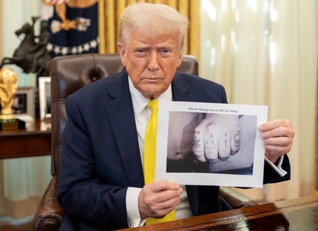 Donald Trump dans le Bureau ovale tenant une feuille imprimée avec une photo de tatouage et le nom « Kilmar Abrego Garcia MS-13 Tattoo ». 
Sur la photo on voit un poing fermé. Sur 4 doigts de gauche à droite, une feuille de cannabis, un smiley, une croix chrétienne, une tête de mort. 