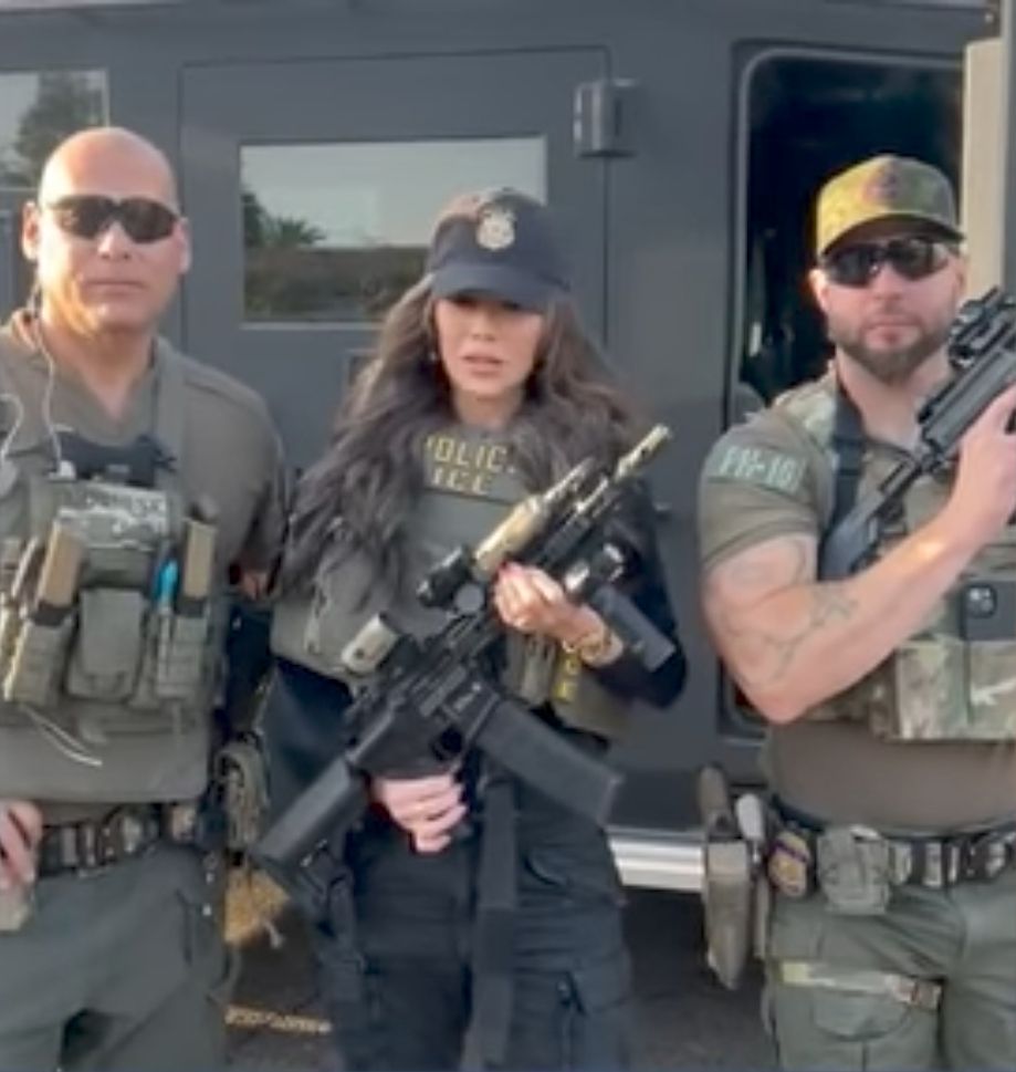 Kristi Noem pose au centre, en tenue tactique, fusil d’assaut en main, entourée de deux agents en uniforme militaire. Tous trois sont armés et en équipement lourd, debout devant un véhicule blindé
