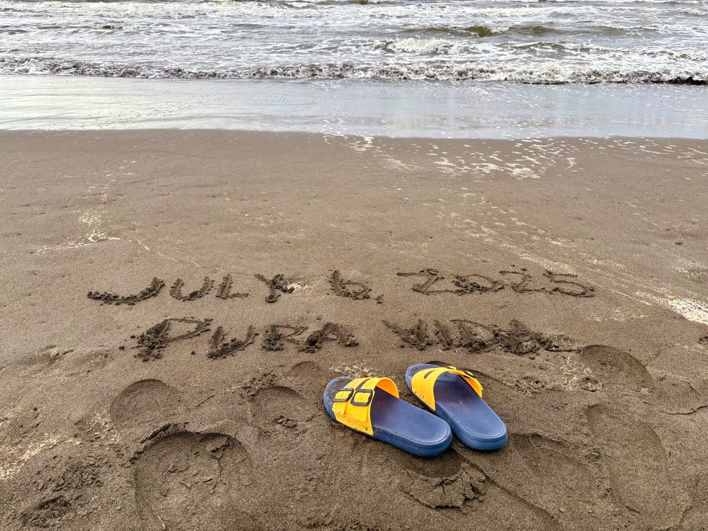 KP’s sandals on the beach in Costa Rica with “July 6, 2025” and “Pura Vida” written in the sand. Waves in the background. 