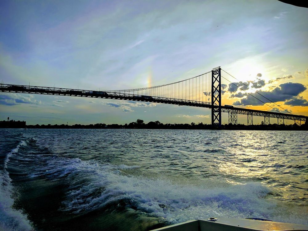 A view from a boat moving through water, with waves in the foreground created by the boat’s motion. Above, the ambassador bridge with a dark silhouette spans the scene, its cables and towers illuminated by the soft light of a setting or rising sun. The sky is a mix of blue and golden hues with scattered clouds, and a faint rainbow-like arc is visible near the sun. In the distance, a shoreline with trees and structures is faintly outlined.