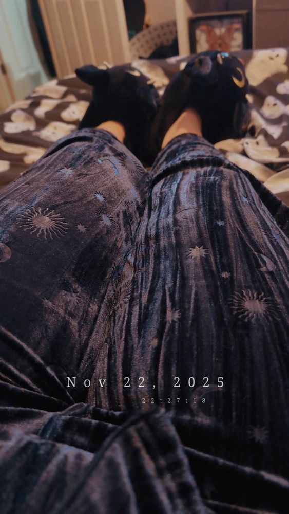 Photo of my legs outstretched on top of my bed. I'm wearing black slippers shaped like giant bats and soft dark blue pyjamas with a sun and moon pattern. A date and time stamp is on the bottom of the photo - Nov 22, 2025 @ 22:27