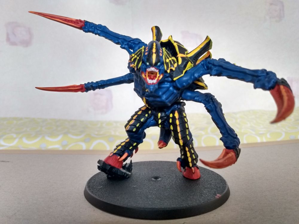 A model of a Screamer-Killer Carnifex, painted with dark-blue exoskeleton, black-and-yellow armour, red talons and hooves, and green eyes, with some bright orange highlights.