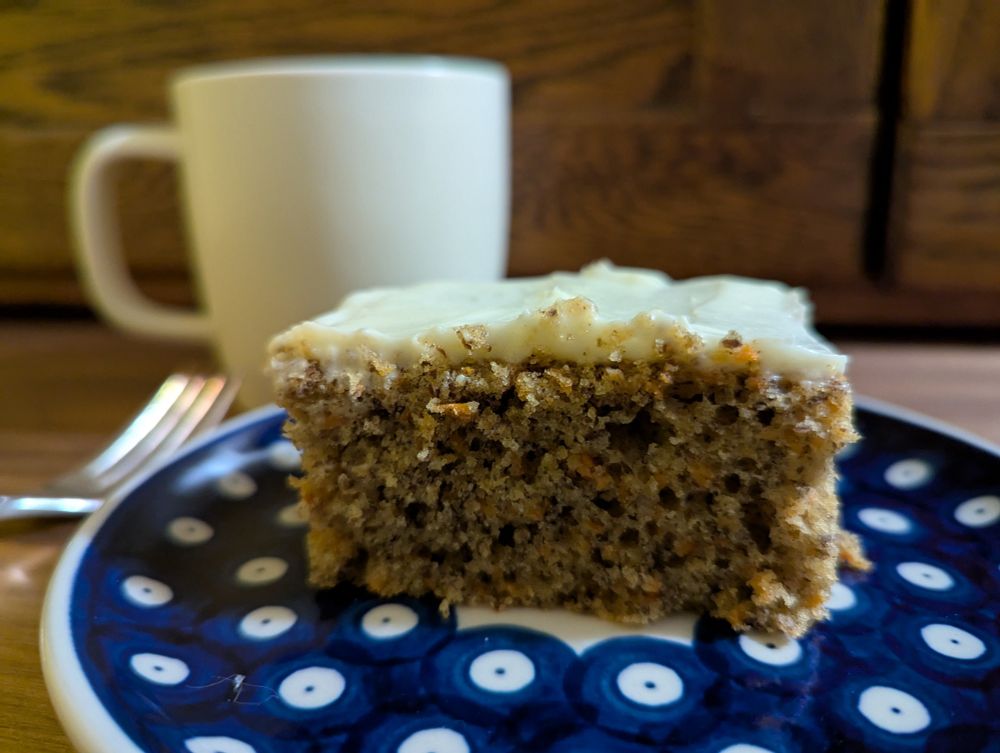 Slice of carrot cake on a blue and white plate with a cup of coffee and a fork in the background.